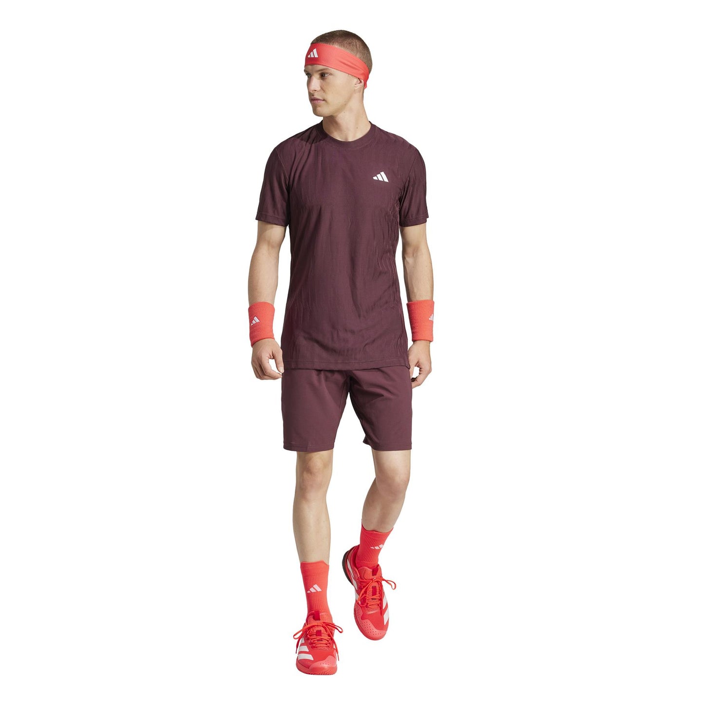 Dressed in maroon athletic wear with red accessories like a headband and HEAT.RDY wristbands, a person strikes a stylish pose against a white backdrop. The lightweight ensemble includes adidas Ergo Men's Padel Shorts - Ruby, showcasing the iconic sports logo for added performance.