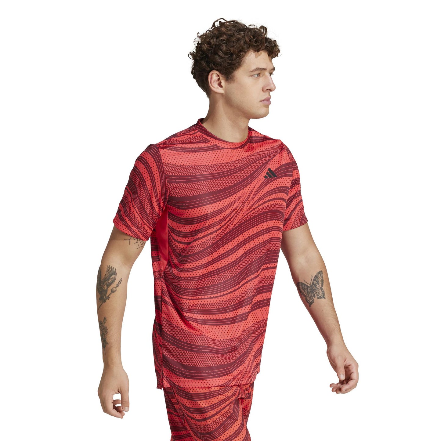 A tattooed, curly-haired man wears a red ADIDAS Mens Club Graphic Padel T-Shirt made of recycled materials. Featuring AEROREADY technology and a small chest logo, he stands looking left.