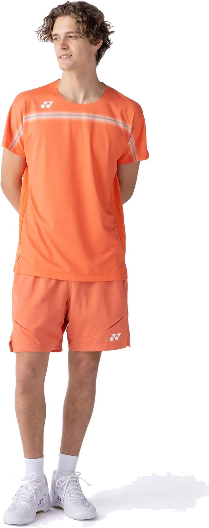 A young person stands with hands behind their back, wearing a Yonex 10711YX Crew Neck Mens Padel T-Shirt in Cyber Orange and matching shorts, plus white socks and sneakers, against a white background.