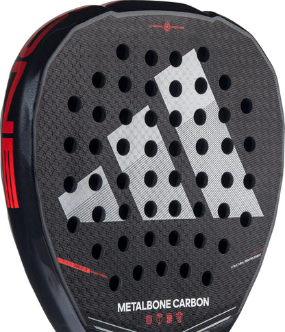 Close-up of the adidas Metalbone Carbon 2026 padel racket, showing a honeycomb pattern, bold white geometric logo, circular holes, red accents, and "METALBONE CARBON" text—ideal for advanced players who value precision.