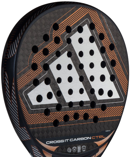 Close-up of the adidas Cross It Carbon Ctrl 2026 Padel Racket – Black, featuring a white geometric design with orange accents. This carbon fibre racket offers an Extra Power Grip and has multiple circular holes on its surface.