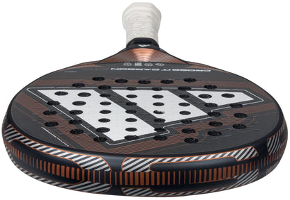 A close-up of the adidas Cross It Carbon Ctrl 2026 Padel Racket - Black, featuring a carbon fibre design with orange accents, white Extra Power Grip, and a distinctive pattern of holes on its face.