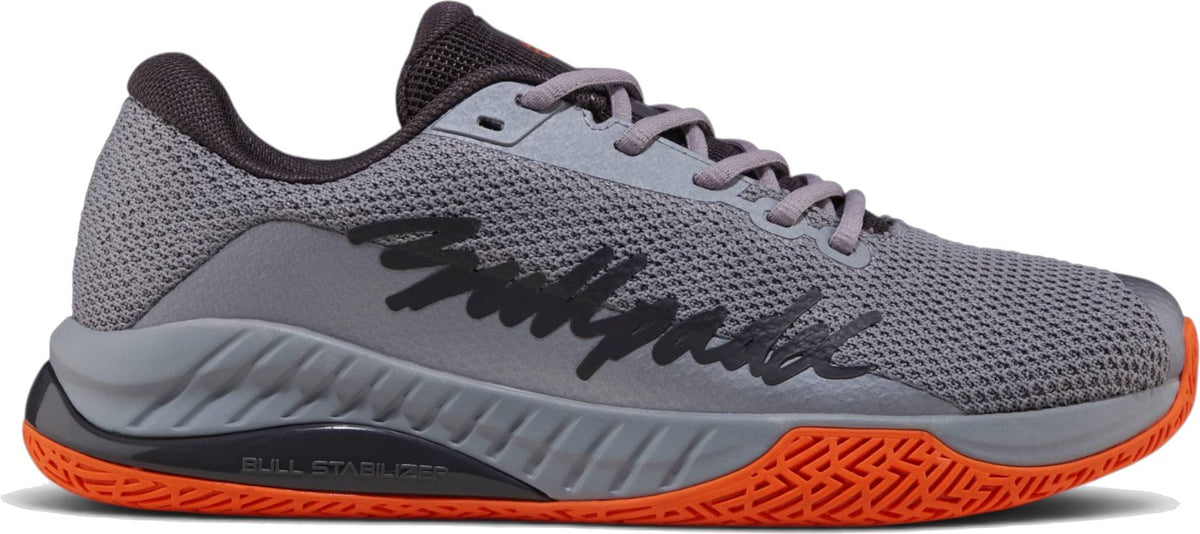 The Bullpadel Ionic 26I Mens Padel Shoes - Dark Grey feature black “Bullpadel” lettering, an orange textured sole, and a mesh upper—ideal for intermediate to advanced players for sport or casual wear.
