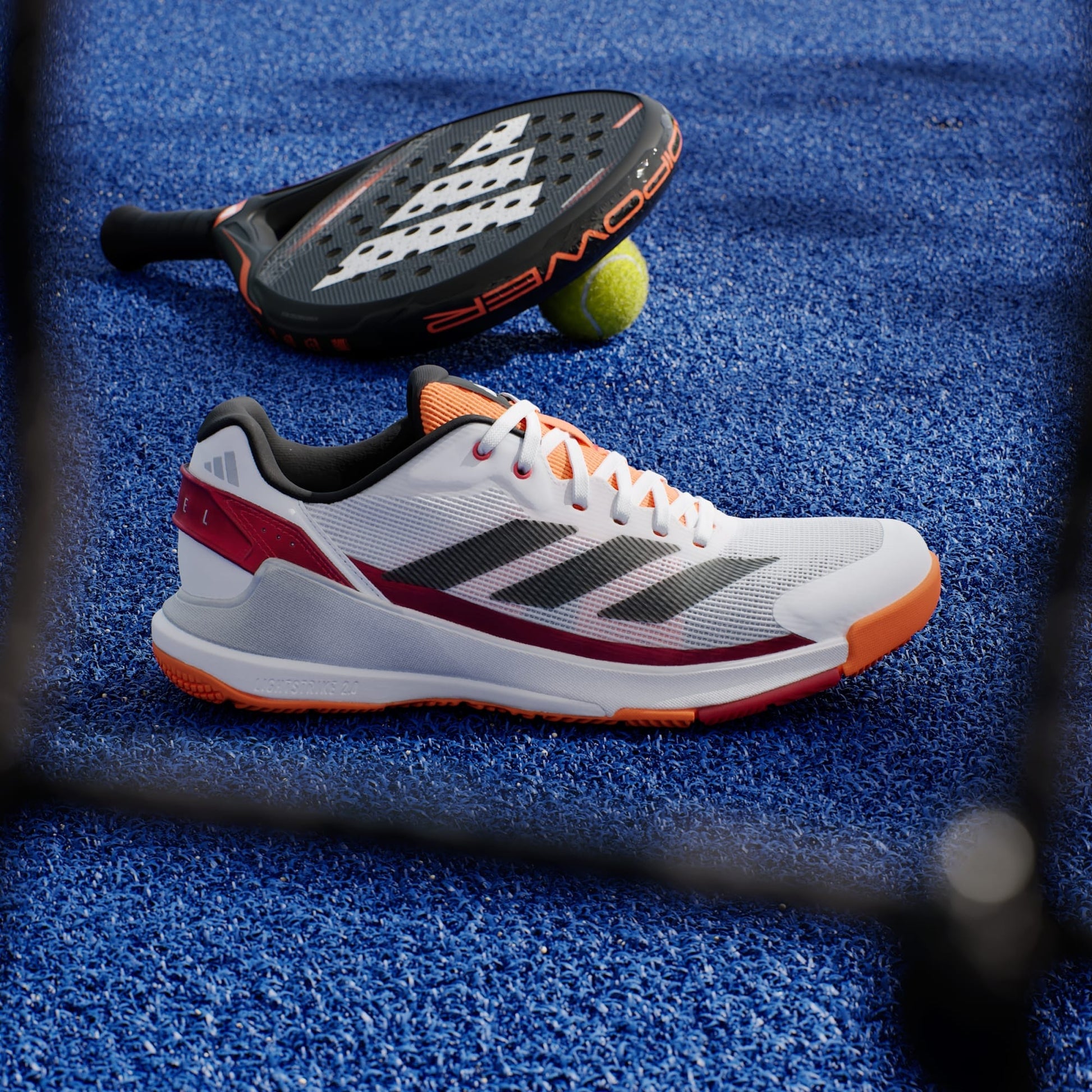 An adidas Crazyquick LS Men's Padel Shoe in white and orange rests on a blue court, beside a black paddle racket and a yellow tennis ball.