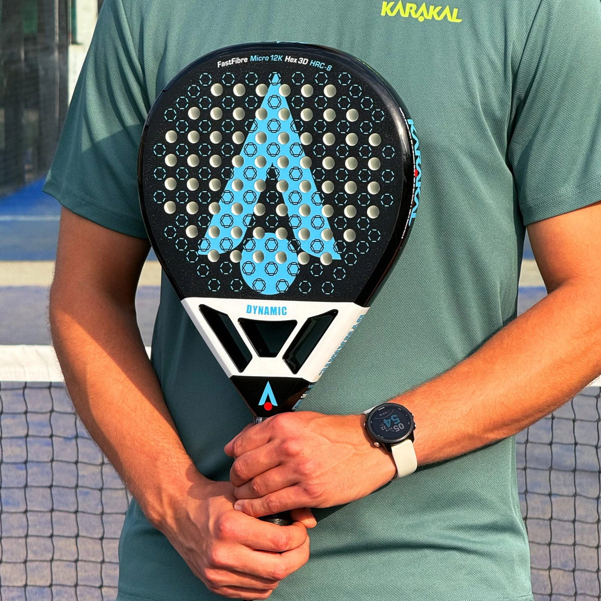 A person in a green t-shirt holds a black and blue Karakal Chaos Dynamic 2026 padel racket with a teardrop-shaped head and a perforated surface.