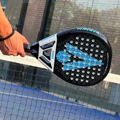 A hand holds a black and white Karakal Chaos Dynamic 2026 padel racket with a blue geometric pattern on its face, positioned in front of a court fence.