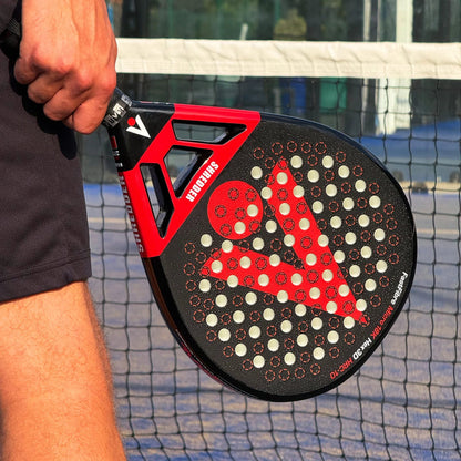 A person holds a black and red Karakal Chaos Shredder 2026 padel racket, featuring a perforated 18K carbon graphite surface, in front of a court net.