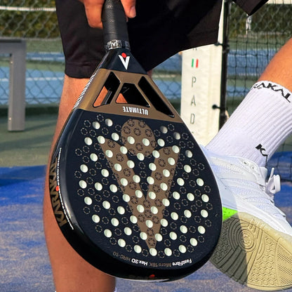 A person holds a black and bronze Karakal Chaos Ultimate 2026 padel racket, featuring a teardrop shape and a textured, hole-punched surface.