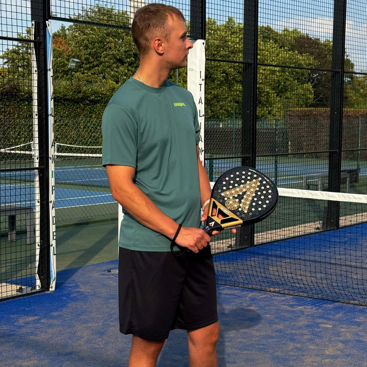 A man in a green t-shirt stands on a blue padel court holding a black and gold Karakal Chaos Ultimate 2026 padel racket.