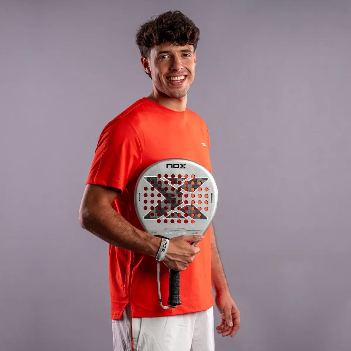 A smiling man in a bright orange t-shirt holds a silver and black NOX AT10 Genius 12K Alum Xtrem By Agustín Tapia 2027 padel racket.