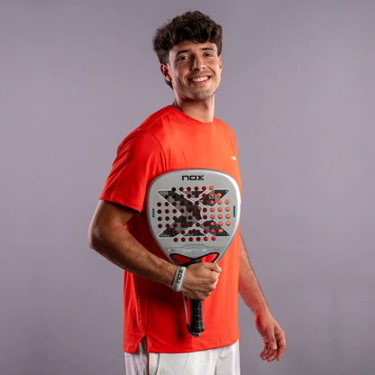 A smiling man in a red t-shirt holding a silver NOX AT10 Genius Attack 12K Alum Xtrem By Agustín Tapia 2027 padel racket with a black and red patterned center.