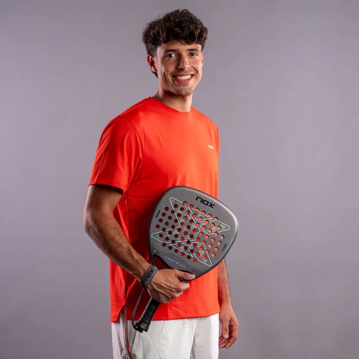 A smiling man in an orange t-shirt holding a NOX AT10 Genius Attack 18K Alum By Agustín Tapia 2027 padel racket.