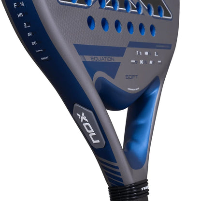 Close-up of a grey and blue NOX Equation Soft Advanced 2027 padel racket with a perforated face, showing the handle, bridge, and technical feature labels printed on the frame.