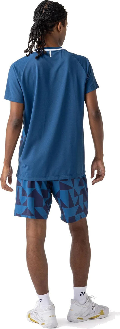 A person with braided hair faces away, wearing the Yonex 10712YX Crew Neck Men's Padel T-Shirt in Indigo Blue, blue geometric shorts, white socks, and white sneakers against a white background.