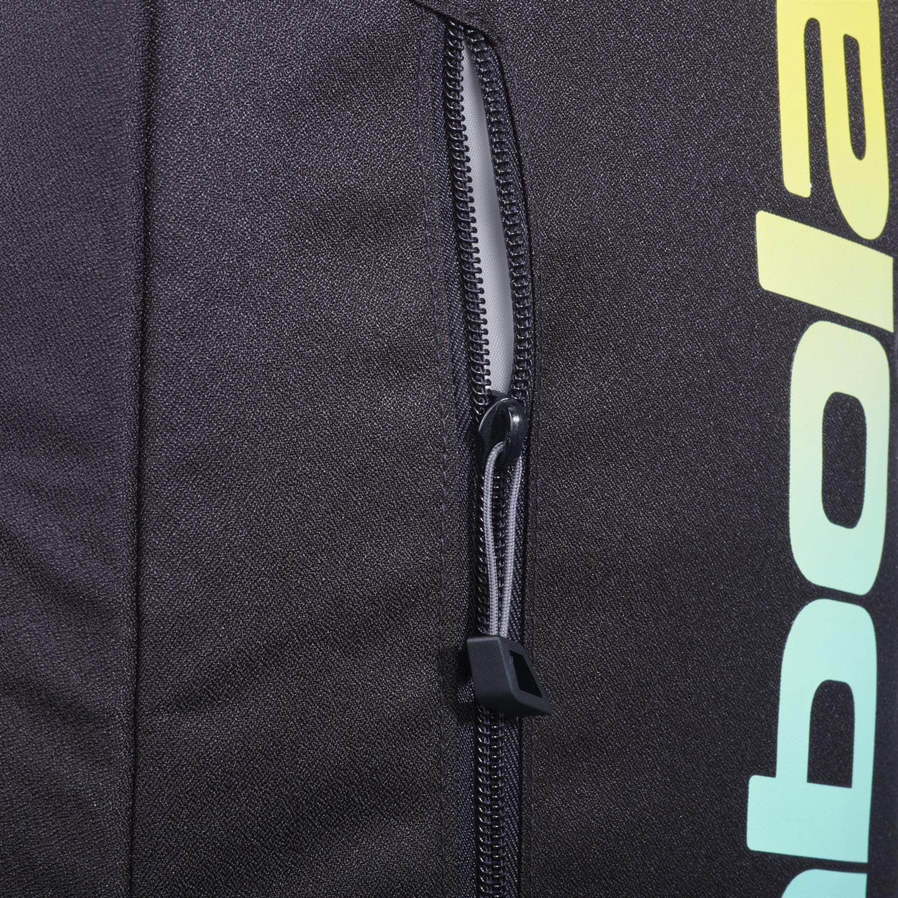 Close-up of the black zippered pocket on the Babolat Court Lite Padel Backpack - Black / Multi, showing part of the "Babolat" logo in a yellow-green gradient and a peek at the dedicated racket compartment on the right.