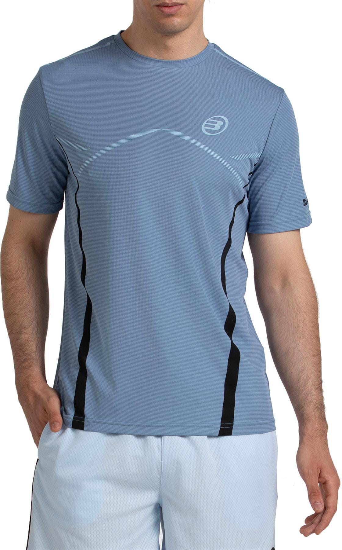 A man in a Bullpadel Cabore Men's Padel T-Shirt in Shadow Blue with Quickerdry tech, plus white shorts, stands facing the camera with one hand in his pocket against a white background.