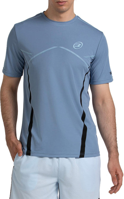 A man in a Bullpadel Cabore Men's Padel T-Shirt in Shadow Blue with Quickerdry tech, plus white shorts, stands facing the camera with one hand in his pocket against a white background.