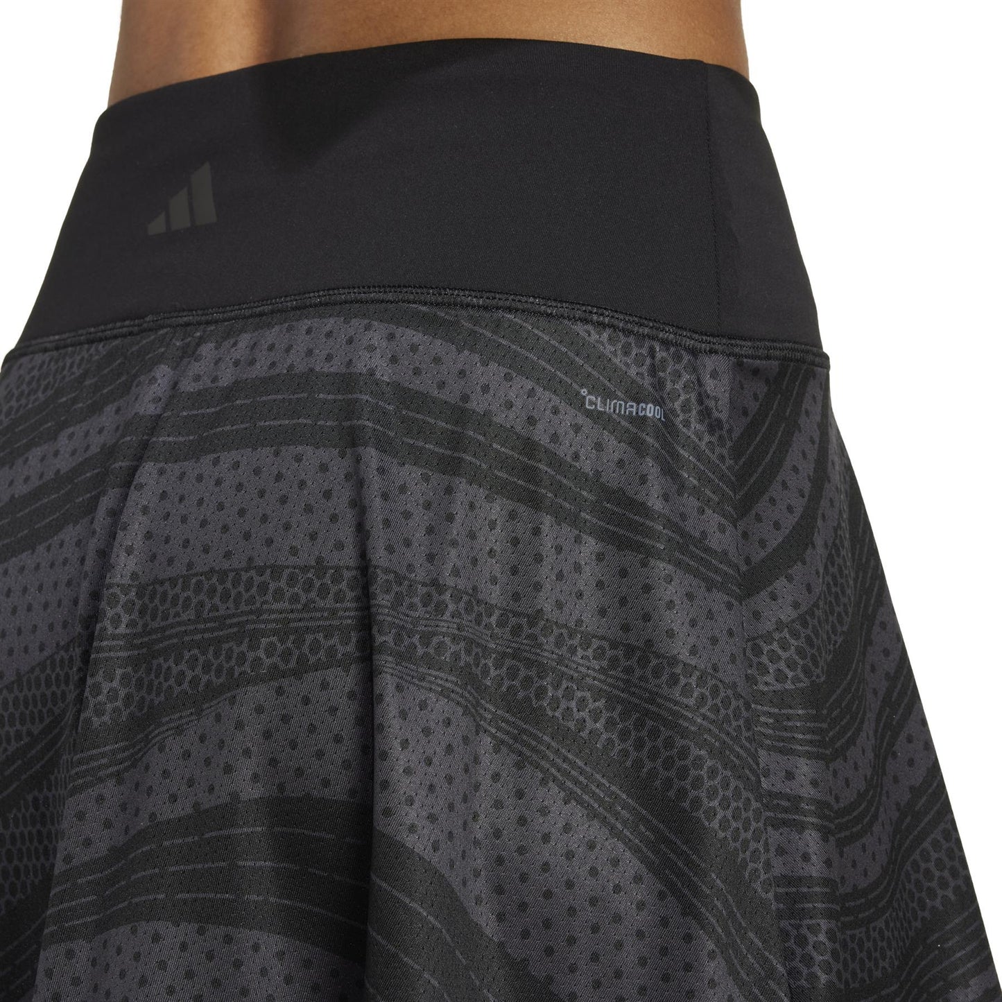 Close-up of a person's waist in the adidas Women's Club Graphic Padel Skirt - Black, highlighting its subtle geometric pattern. The skirt features "Climacool" text with the brand logo, emphasizing comfort and breathability from recycled materials.