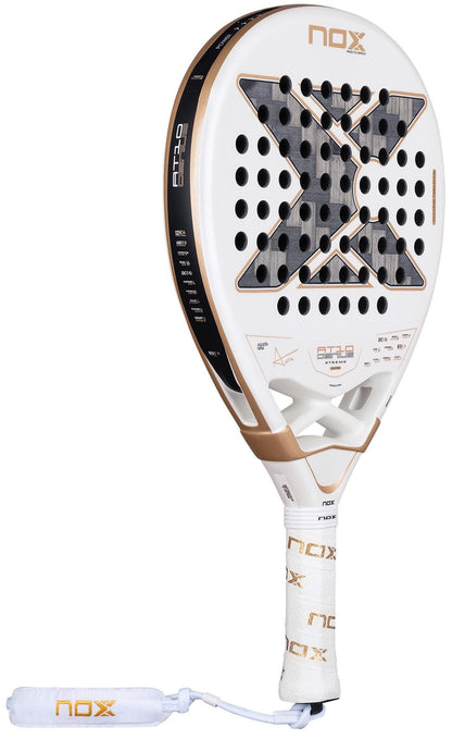 The NOX AT10 Genius 12K Alum Extrem By Agustín Tapia 2026 Padel Racket features a white and gold design, bold black X on the face, multiple perforations, and a handle wrapped with the NOX logo.