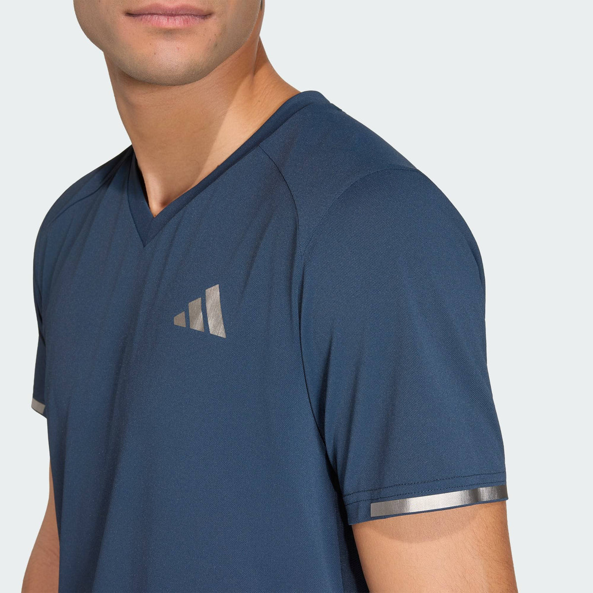 A person wearing a blue ADIDAS Climacool+ Freelift Pro Mens Padel T-Shirt - Aurora Ink with metallic sleeve stripes and a silver geometric chest logo, shown from the shoulders up.
