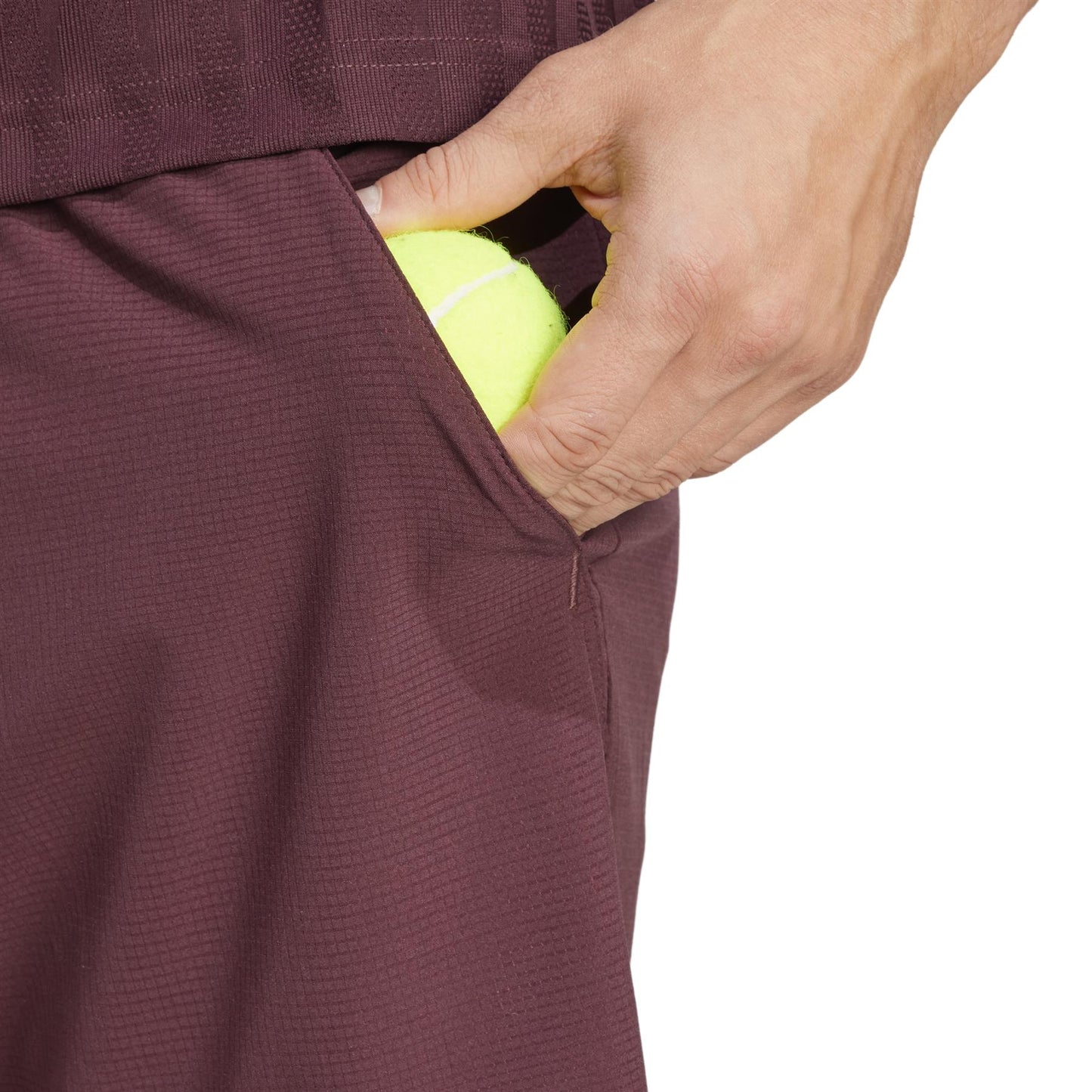 A hand places a yellow tennis ball into the pocket of ADIDAS Ergo Mens Padel Shorts in Ruby. The focus is on the hand, emphasizing the lightweight HEAT.RDY fabric against a plain white background.