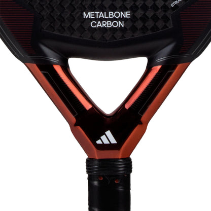 Close-up of the ADIDAS Metalbone Carbon 3.3 Padel Racket, in black and copper, on a textured surface. The frame has a glossy metallic finish with a triangular throat, featuring the adidas logo at the base and crafted with premium 6K Carbon for enhanced performance.