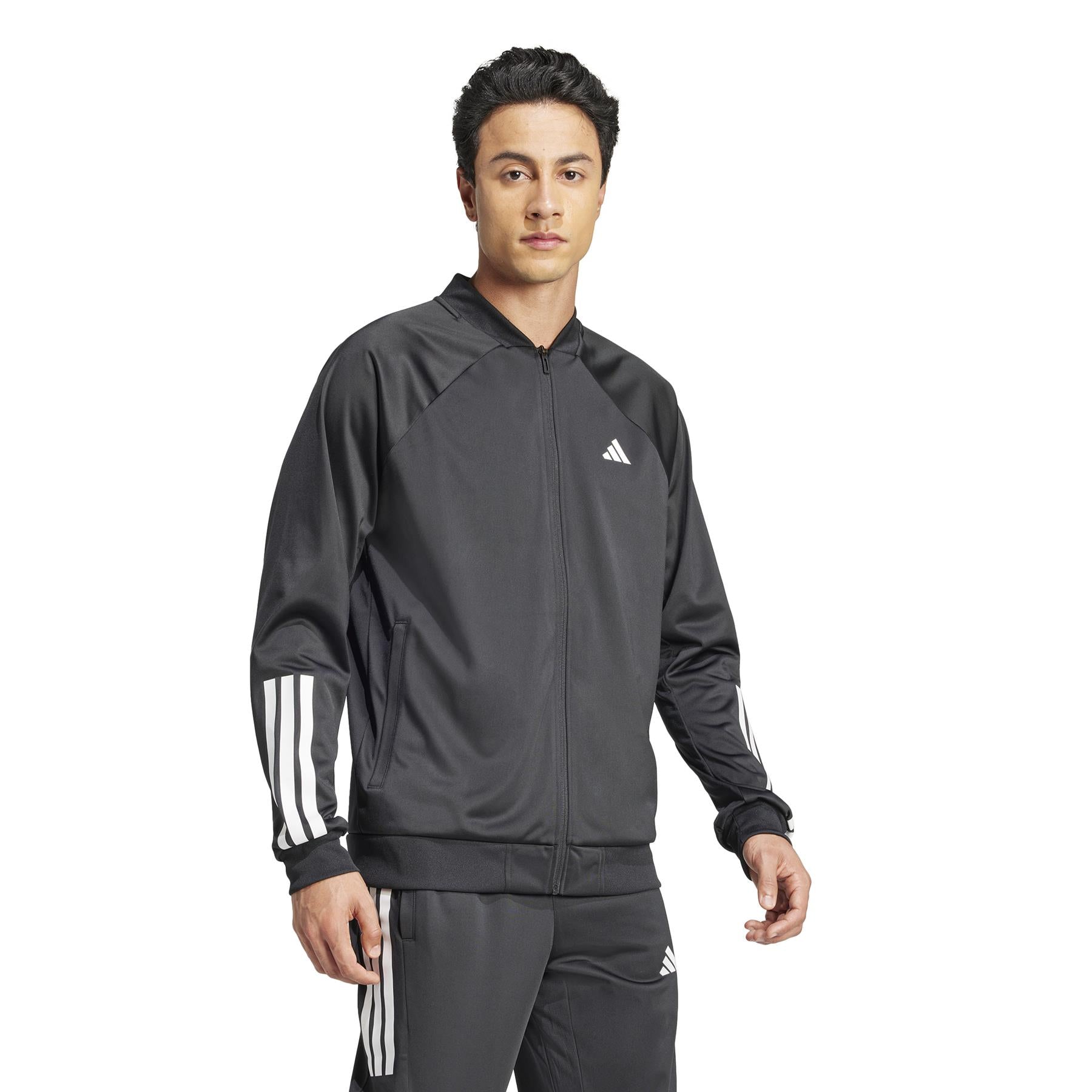 A person stands confidently in an adidas Men's 3 Stripe Knit Padel Jacket - Black, featuring white stripes and a subtle logo. Paired with matching pants, their short dark hair and forward gaze create a striking pose against a plain white backdrop.