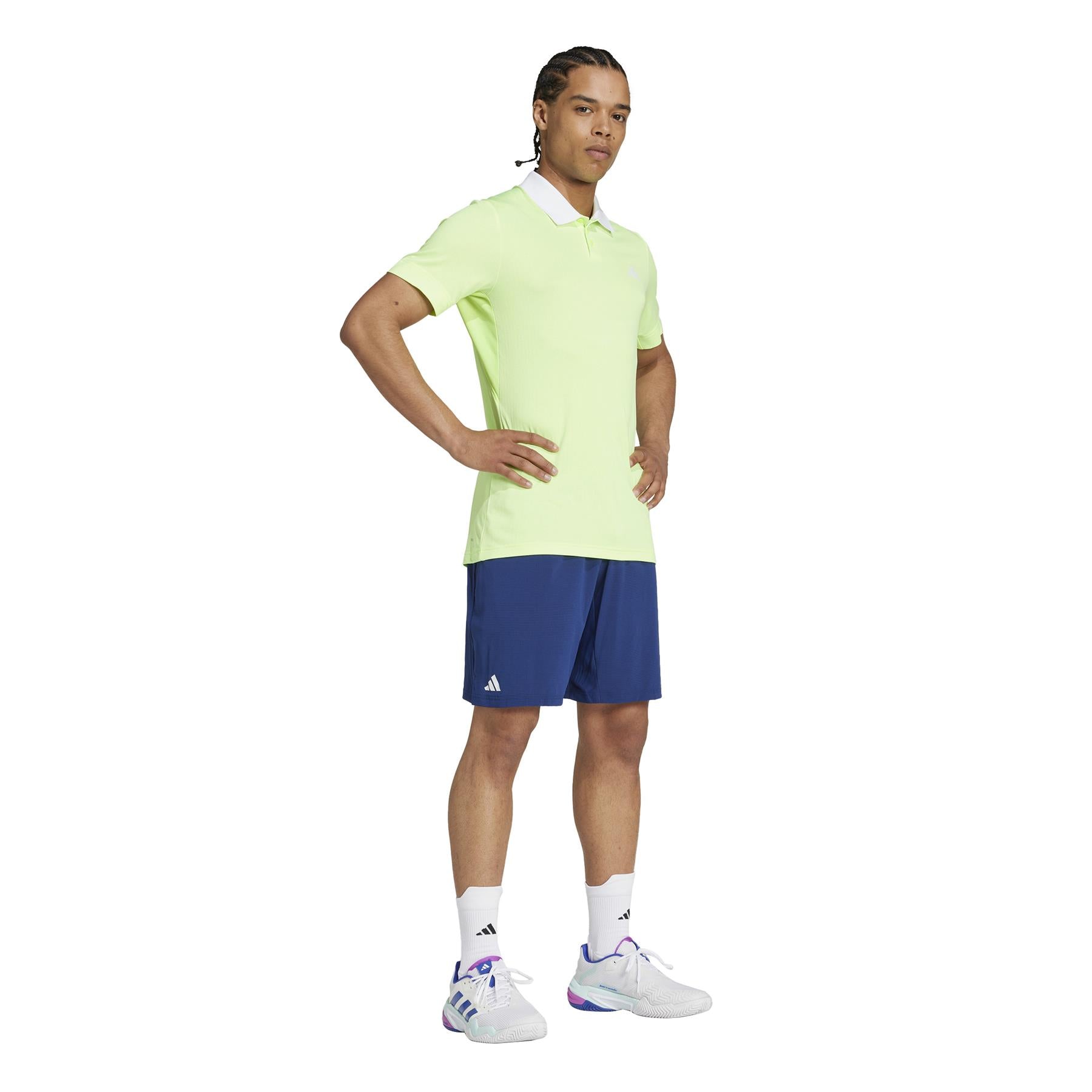 A person stands confidently with crossed arms against a plain white background, wearing a light green polo shirt, ADIDAS Ergo Men's Padel Shorts in navy, and lightweight white sneakers with colorful accents stylishly crafted from recycled materials.