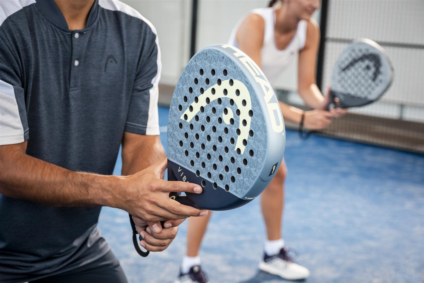 On a blue padel court, two players in sportswear grip their HEAD Vibe 2026 Blue / Yellow Padel Rackets by HEAD, hands poised on the fibreglass surface, anticipation building before the match.