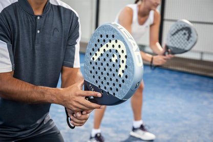 On a blue padel court, two players in sportswear grip their HEAD Vibe 2026 Blue / Yellow Padel Rackets by HEAD, hands poised on the fibreglass surface, anticipation building before the match.