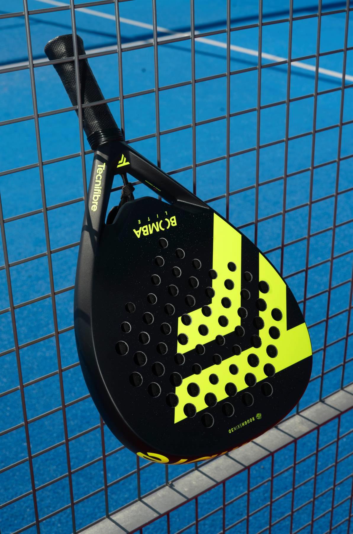 The black and yellow Tecnifibre Bomba Lite Padel Racket by Tecnifibre, featuring circular holes and a fibreglass face, leans against a metal fence with a bright blue padel court in the background.