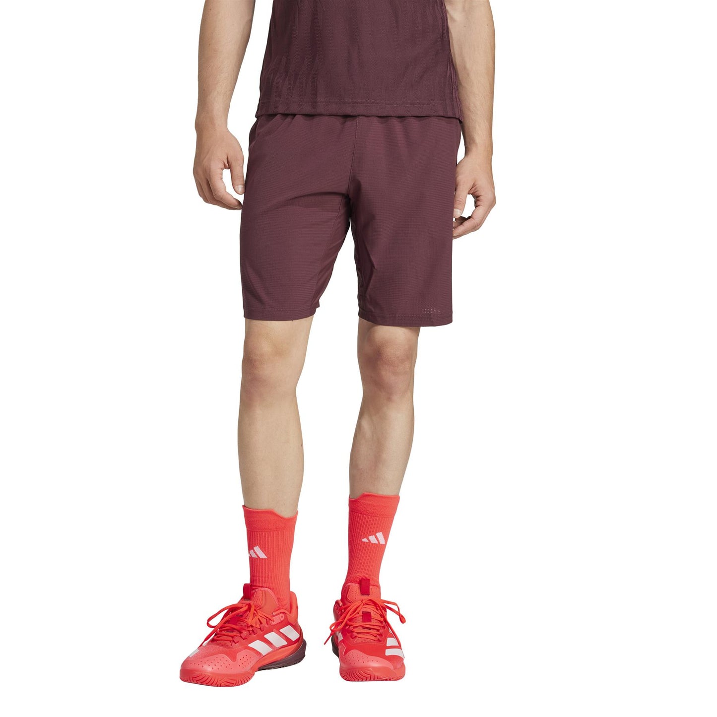 A person in a maroon shirt and ADIDAS Ergo Mens Padel Shorts - Ruby stands against a white background, wearing bright orange socks and matching sneakers with white stripes. The focus is on the clothing from the neck down, highlighting the lightweight shorts made from recycled materials.