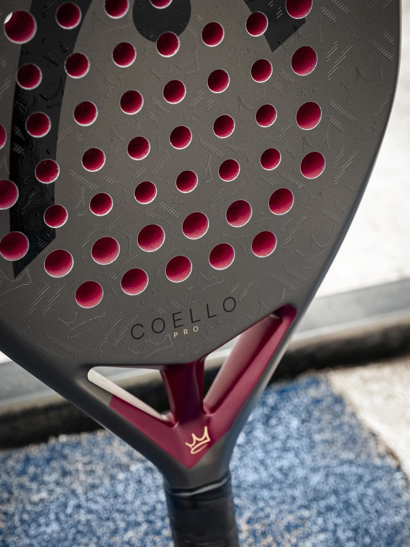 Close-up of a black and maroon HEAD Coello Pro 2026 Padel Racket with round holes and "Coello Pro" on its carbon hybrid surface, resting on a blue textured surface near a window.