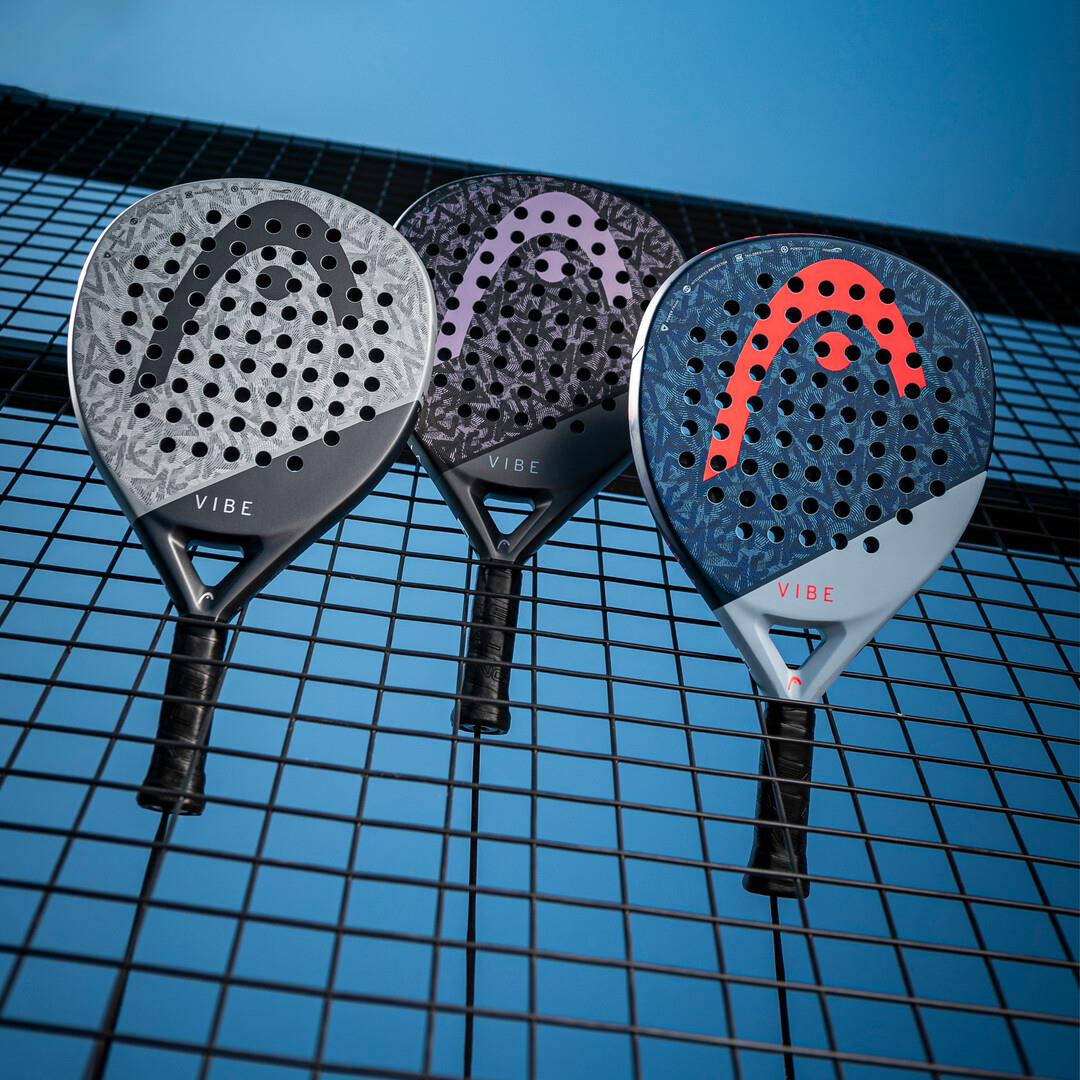 Three HEAD Vibe 2025 padel rackets hang on a wire fence under a clear blue sky. The designs include gray and black, purple accents, and blue with red highlights. Each racket is crafted for easy power and has a fiberglass hitting surface.