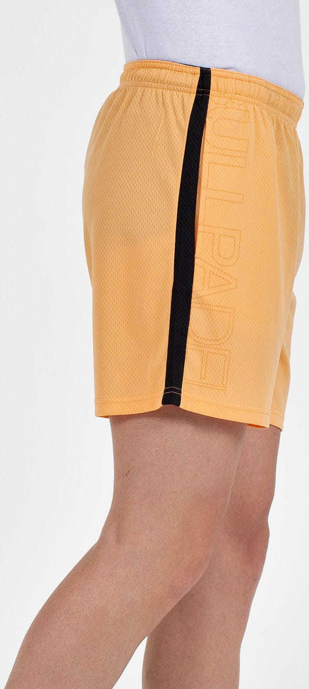 A person wears Bullpadel Campaza Men's Padel Shorts in orange, featuring a black vertical stripe and subtle side text, paired with a white shirt. Only their lower torso and upper legs are visible.