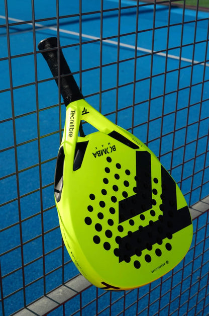 The Tecnifibre Bomba Max Padel Racket, featuring a striking yellow design with black accents and renowned for explosive power, rests against a wire fence on a blue outdoor court.