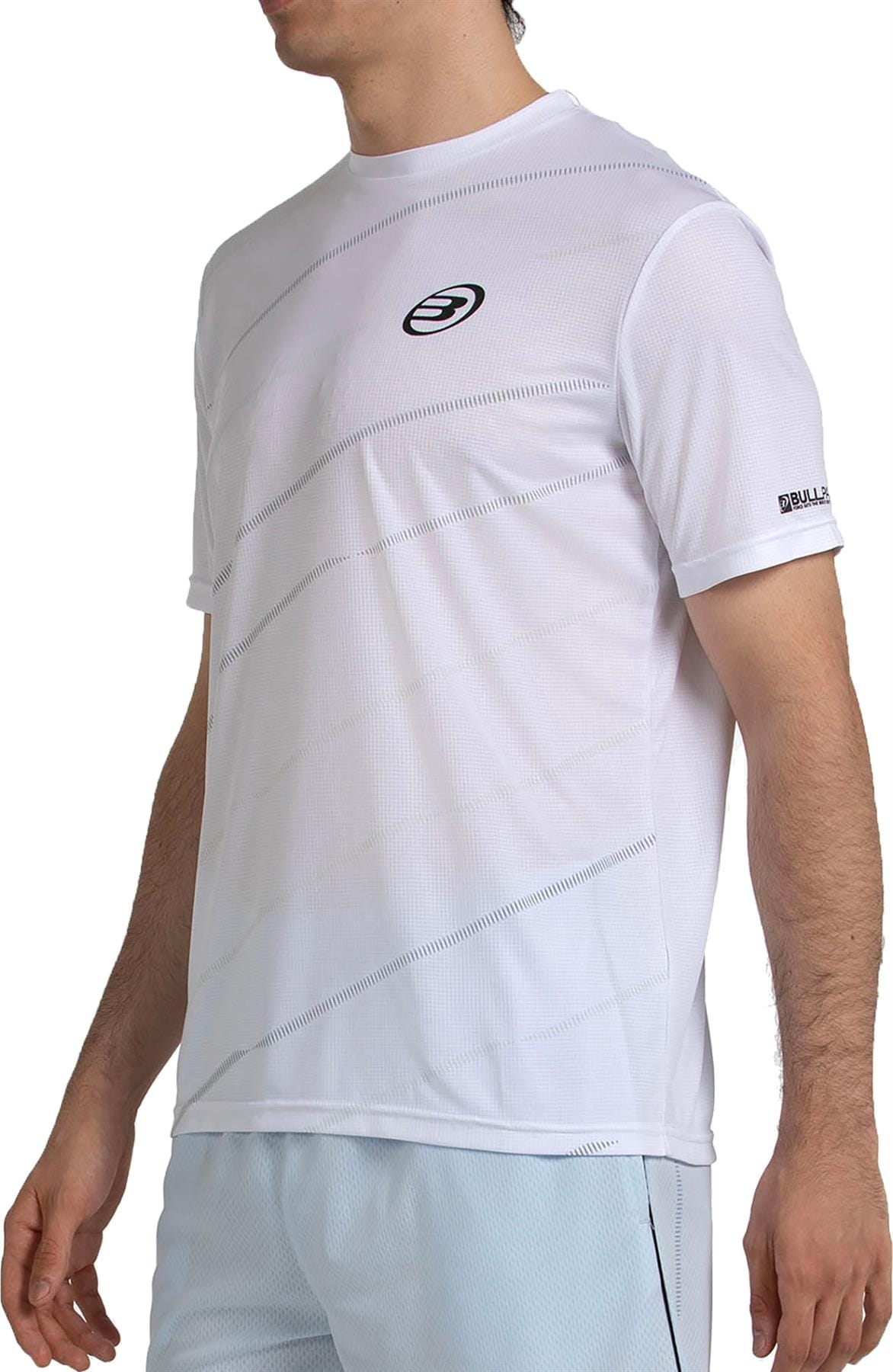 A man wears the Bullpadel Casinas Mens Padel T-Shirt - White, featuring subtle diagonal lines, a small black Bullpadel logo on the chest and sleeve, and quickerdry technology, standing against a plain background.