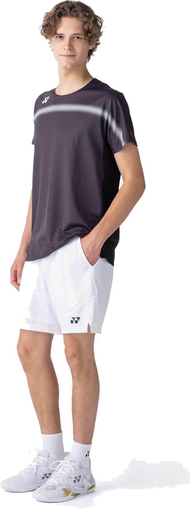A young man with short curly hair and a neutral expression stands with one hand in his pocket, wearing a Yonex 10711YX Crew Neck Mens Padel T-Shirt in black, paired with white shorts, socks, and sneakers. He is facing slightly to the side.