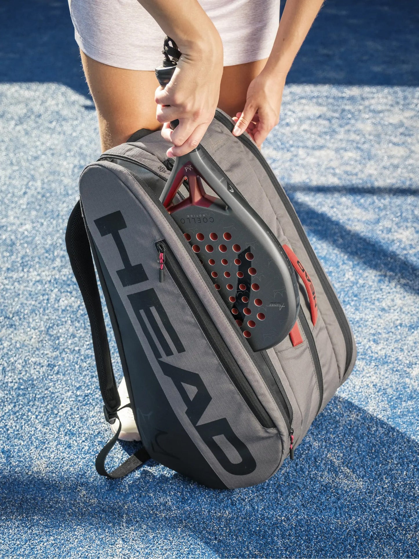 A person puts a black and red padel racket into a spacious HEAD Tour Padel Racket Bag L - Coello by HEAD with CCT+ climate control, on a blue textured court.