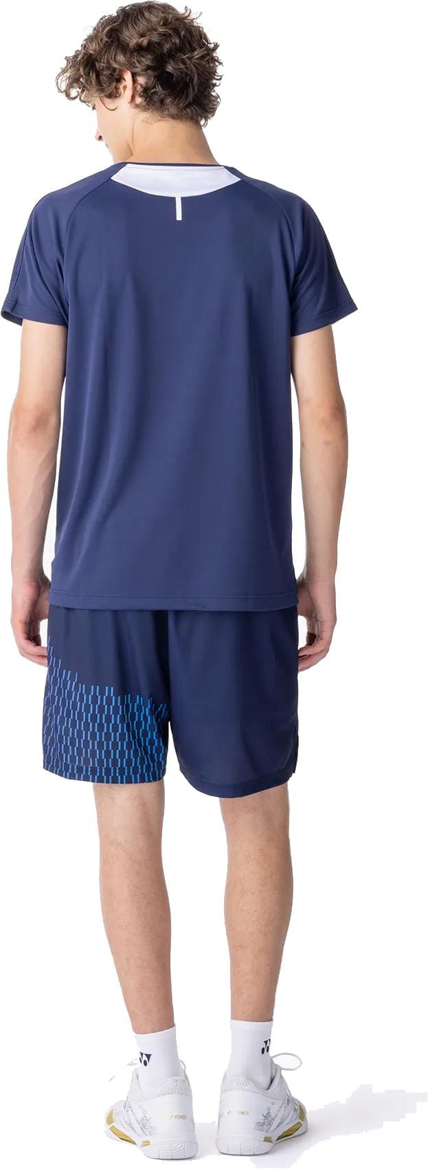 A young man with curly hair stands facing away, wearing the Yonex 10713EX Crew Neck Mens Padel T-Shirt in dark navy, matching blue-patterned shorts, white crew socks, and white athletic shoes.