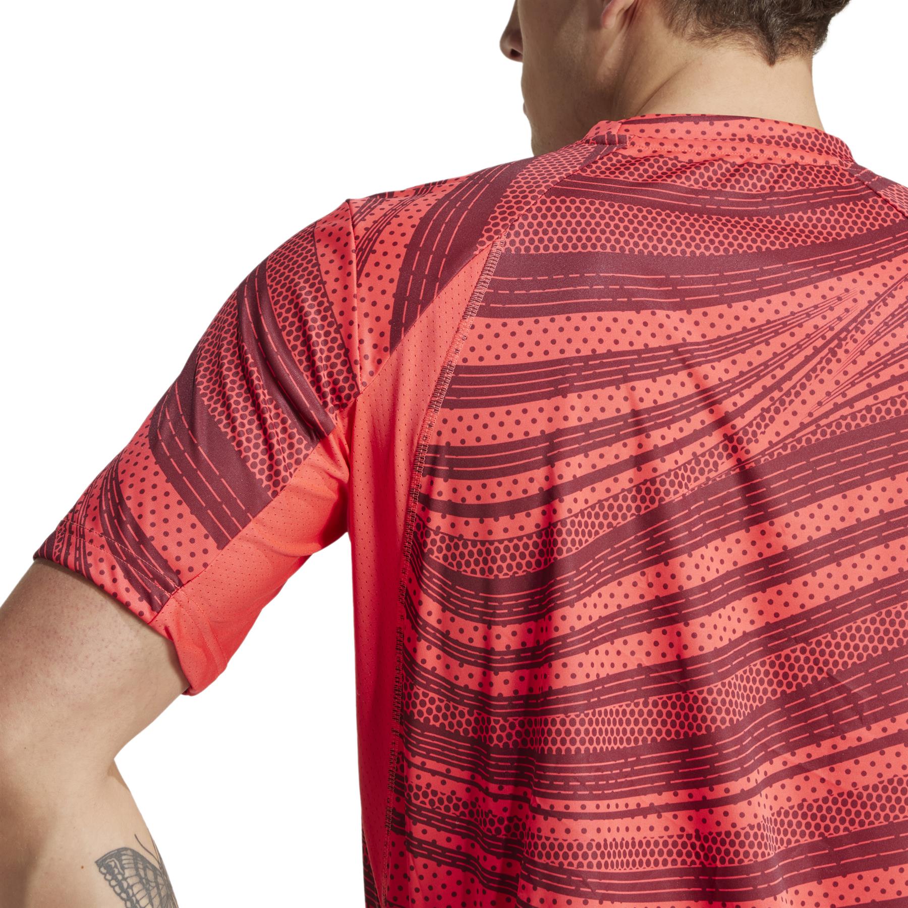 The person wears an adidas Men's Club Graphic Padel T-Shirt in red, featuring dotted and striped designs. Made from recycled materials, this shirt combines style and sustainability. A unique tattoo is visible on the individual's left arm beneath the short sleeves.