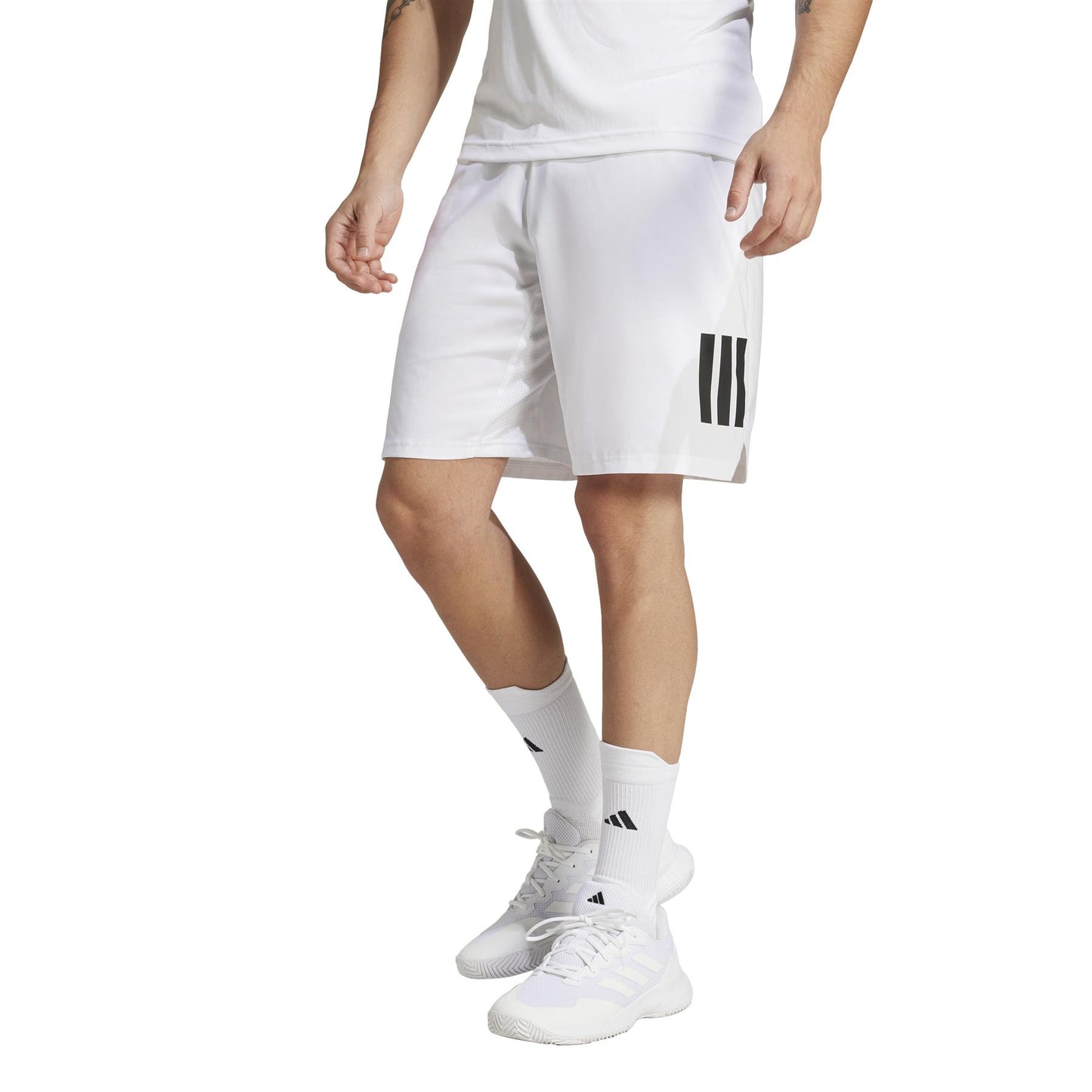 A person is dressed in white athletic attire, featuring adidas Men's 3 Stripe Padel Shorts. The ensemble includes a white shirt, moisture-wicking socks, and sleek sneakers. The focus remains on the lower body against a pristine white backdrop.