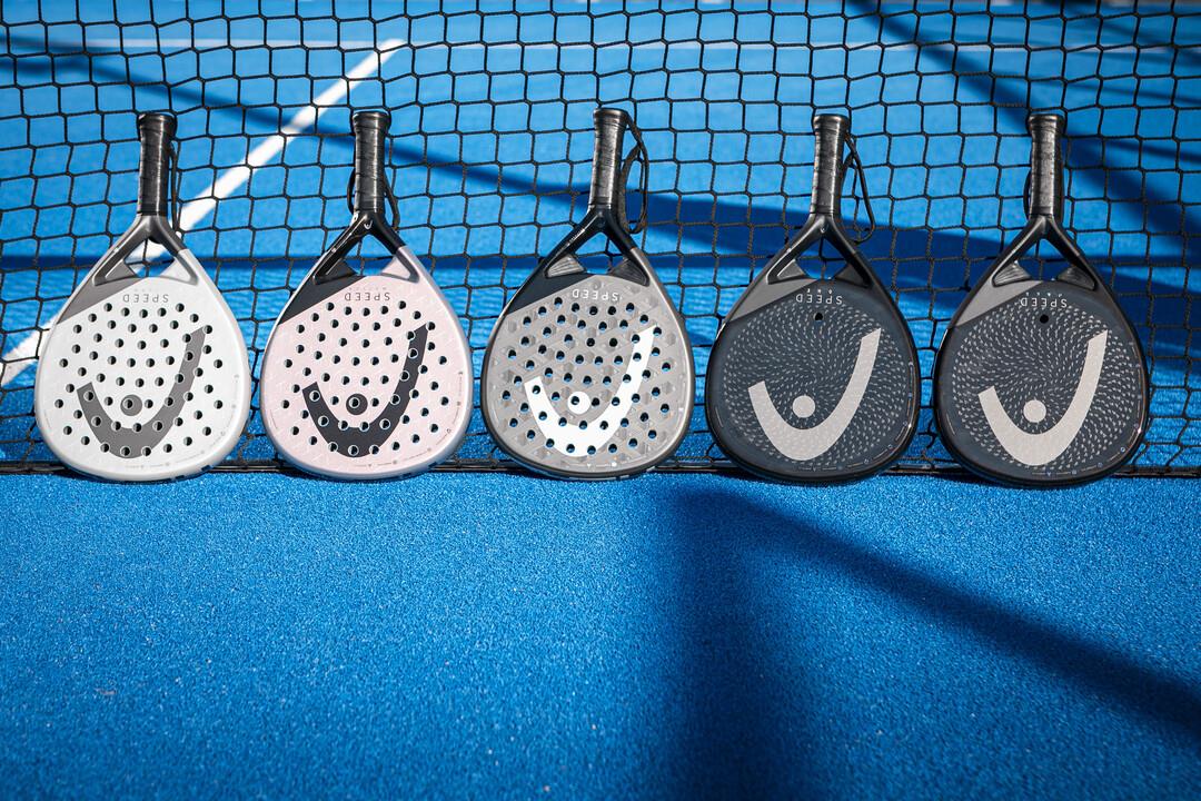 Five HEAD Speed One X 2025 Padel Rackets are propped against a net on a blue court. With black, white, gray, and pink patterns and a curved logo, they feature Extreme Spin technology. Sunlight casts shadows across the surface.