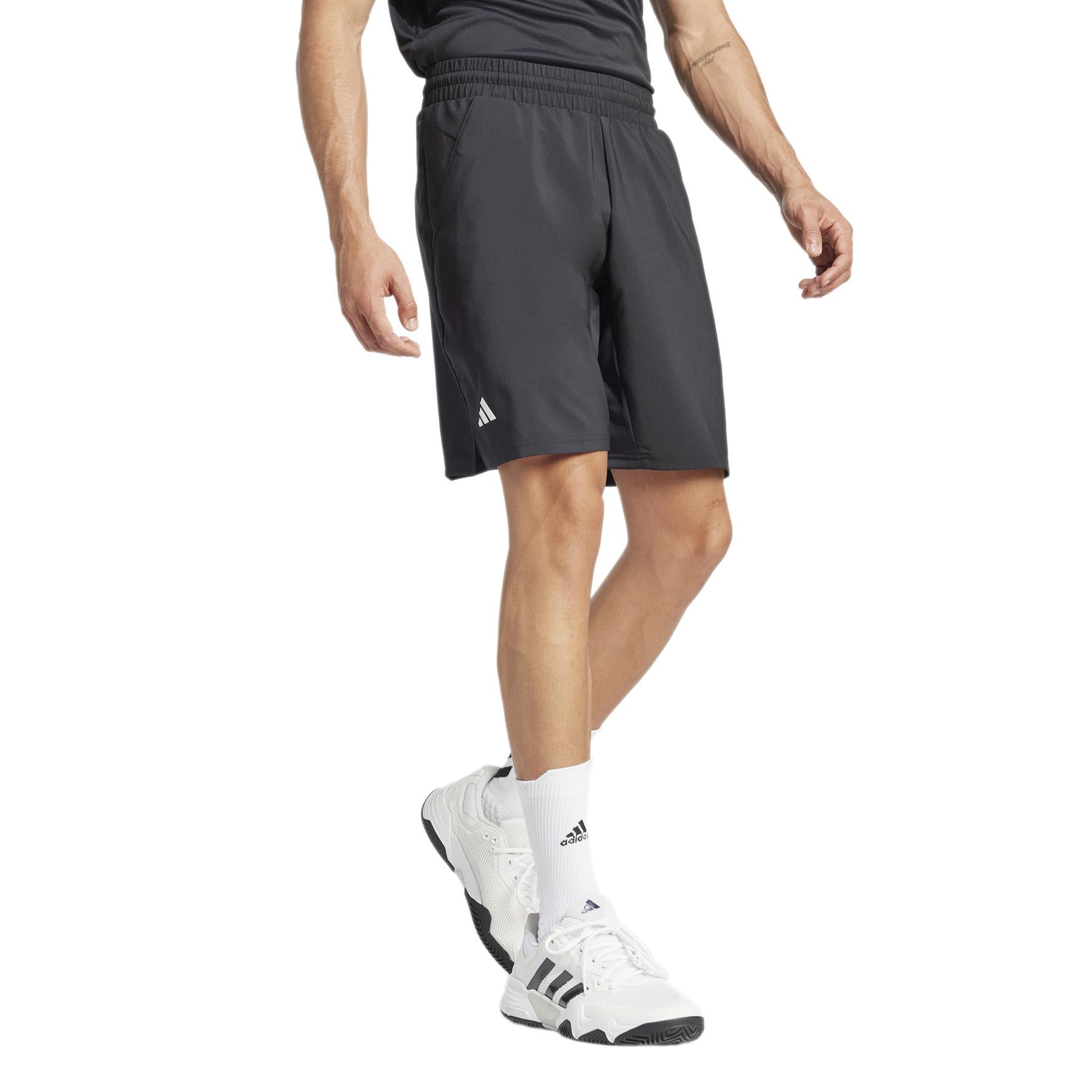 A person wearing ADIDAS Men's 3 Stripe Padel Shorts in black and a matching top stands with one hand by their side, complemented by moisture-wicking white socks and white sneakers featuring black stripes.