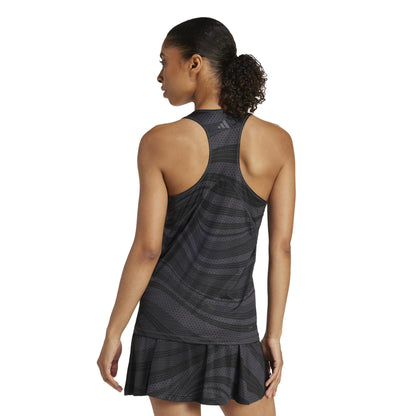 Against a plain white background, a woman with a ponytail wears the ADIDAS Women's Club Graphic Padel Tank Top in black. The dark, sleeveless athletic dress is made from recycled materials and features a subtle striped pattern, reflecting adidas's commitment to sustainability.