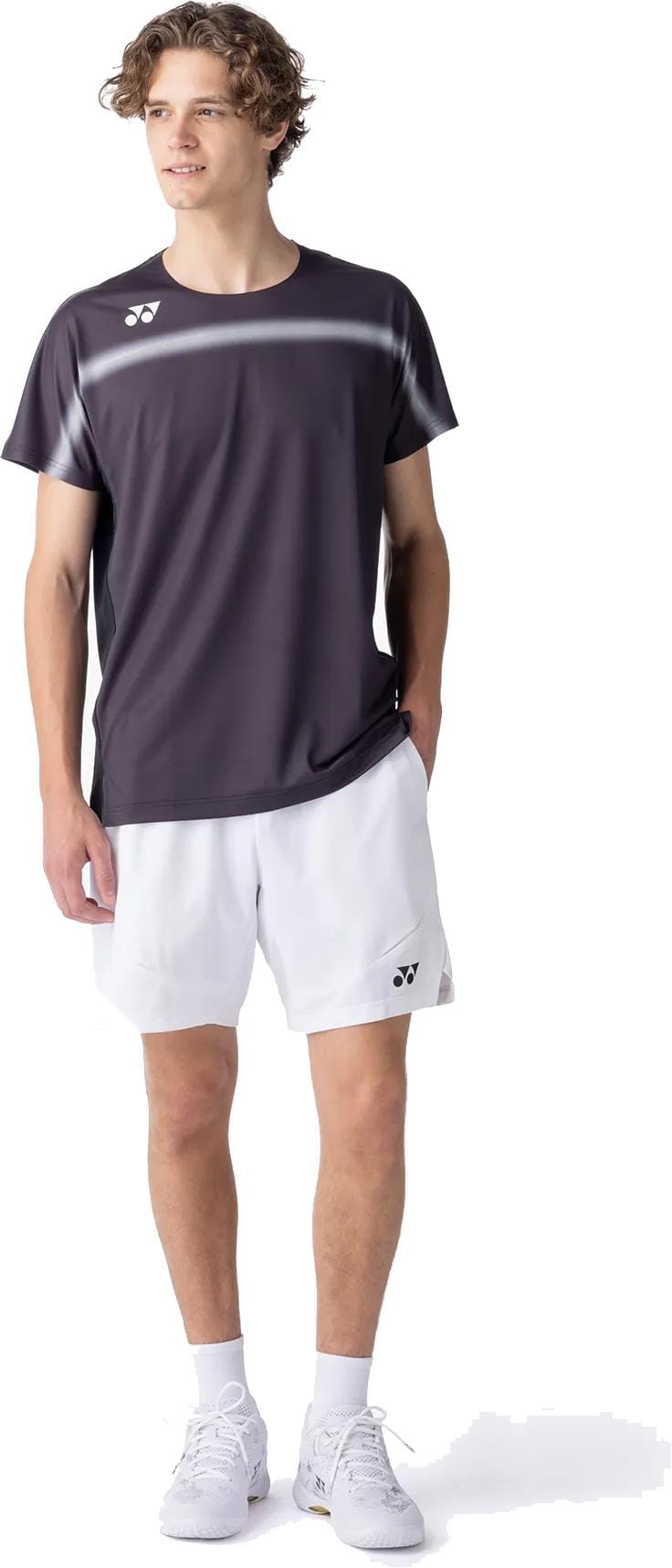 A young man smiles with one hand in his pocket, wearing the Yonex 10711YX Crew Neck Mens Padel T-Shirt in black, paired with white athletic shorts, socks, and sneakers. The Yonex top’s performance fabric keeps him comfortable.