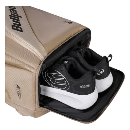 A beige Bullpadel Vertex 25 Padel Backpack - Stone, with a reinforced thermo-lined compartment, holds a pair of black and white athletic shoes. Perfect for carrying your Vertex Padel Racket, it features a side zip and logo.