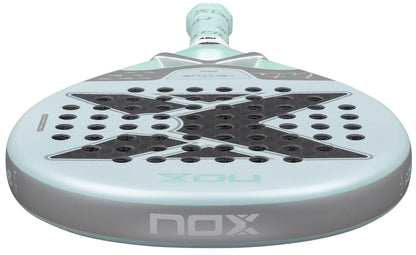 Close-up of the NOX Luxury Ventus 12K Hybrid Lite 2026 Padel Racket in blue, featuring a gray face with a large black "X," small holes on the 12K carbon fibre surface, and a textured grip handle.