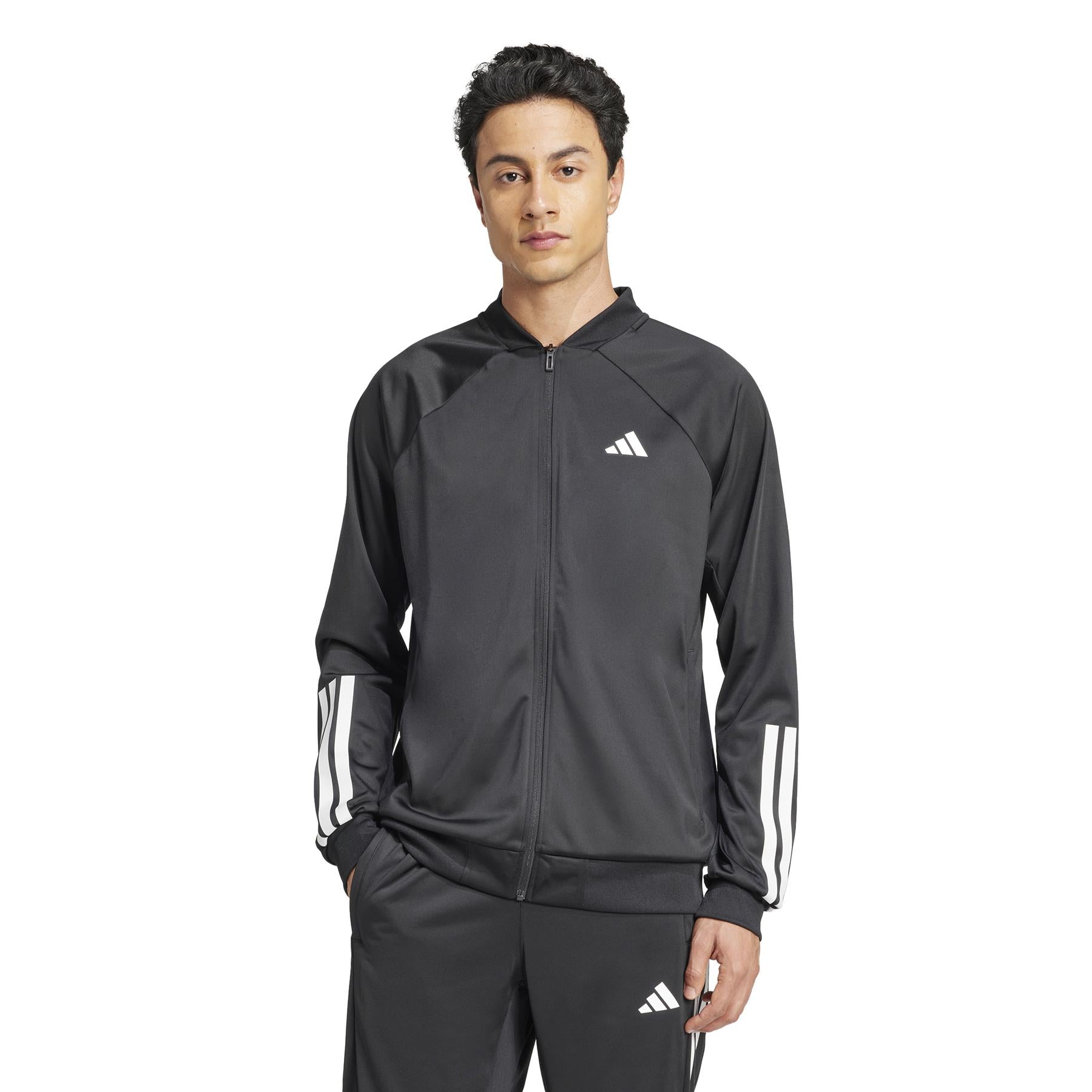 An individual in an ADIDAS Men's 3 Stripe Knit Padel Jacket - Black, featuring white sleeve stripes, stands hands-in-pocket against a plain white background. The outfit includes a zip-up jacket and matching pants from adidas.