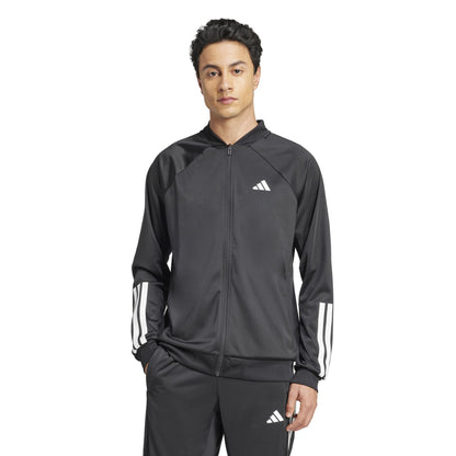 An individual in an ADIDAS Men's 3 Stripe Knit Padel Jacket - Black, featuring white sleeve stripes, stands hands-in-pocket against a plain white background. The outfit includes a zip-up jacket and matching pants from adidas.